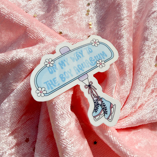 A die-cut sticker featuring a light blue sign that reads 'ON MY WAY TO THE BOY AROMAUM' with small white flowers and an illustration of ice skates, placed on pink glittery fabric.