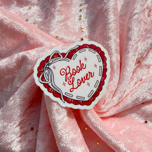 A heart-shaped sticker with the words 'Book Lover' in red script inside a red stitched border, placed on a pink satin fabric background with small gold star accents.