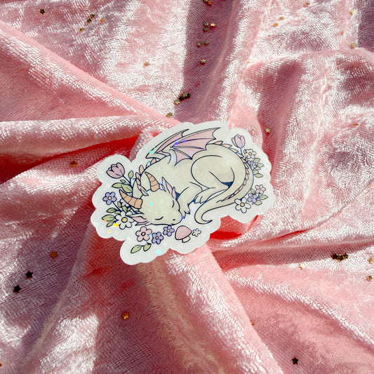 A die-cut sticker featuring a sleeping white dragon surrounded by pastel flowers, placed on a draped pink fabric background with scattered gold glitter.