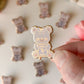 Gold bear-shaped pin with a purple label held by a hand against a background of similar pins.