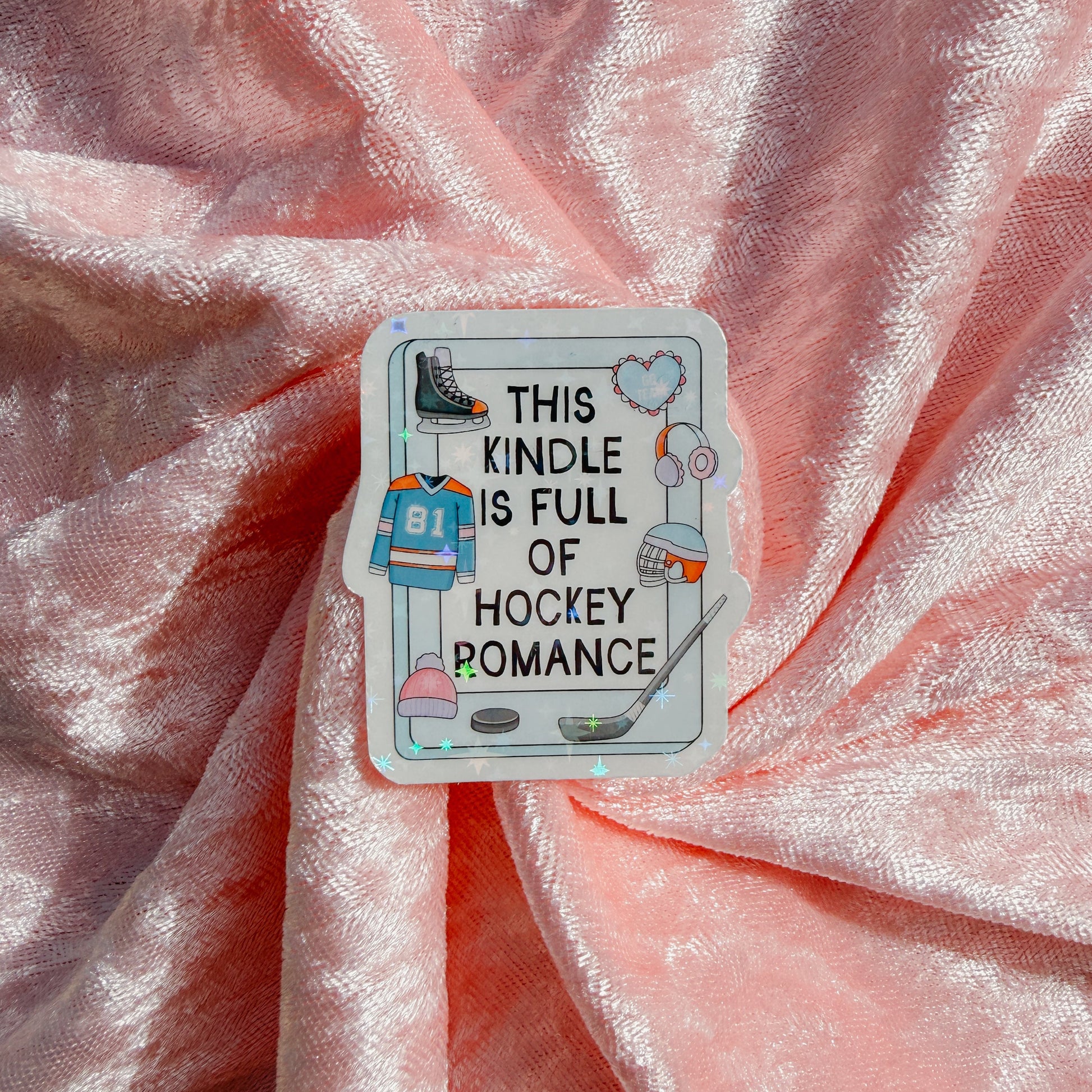 A rectangular enamel pin with rounded corners featuring hockey-themed illustrations and the text 'THIS KINDLE IS FULL OF HOCKEY ROMANCE' in black lettering, placed on draped pink satin fabric.