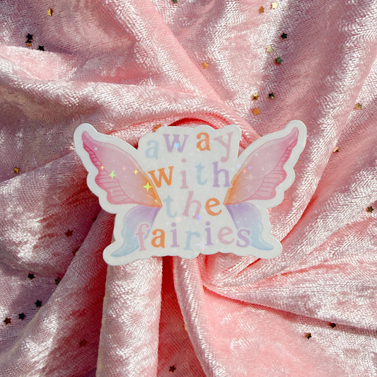 A butterfly-shaped sticker with the words 'away with the fairies' in pastel rainbow letters, placed on a shiny pink fabric background with scattered gold star confetti.