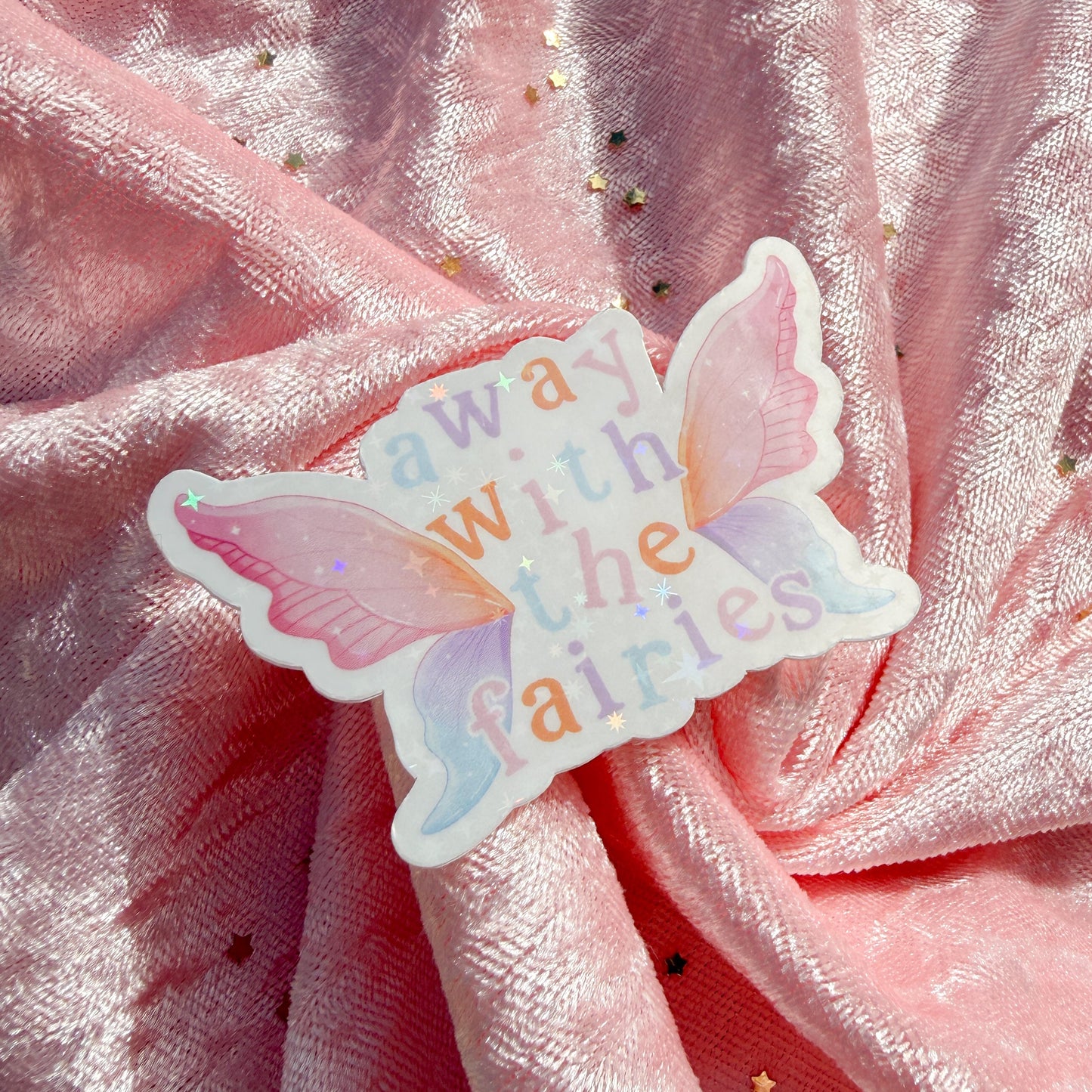 A butterfly-shaped sticker with the words 'away with the fairies' in pastel colors, placed on a draped pink glittery fabric background.