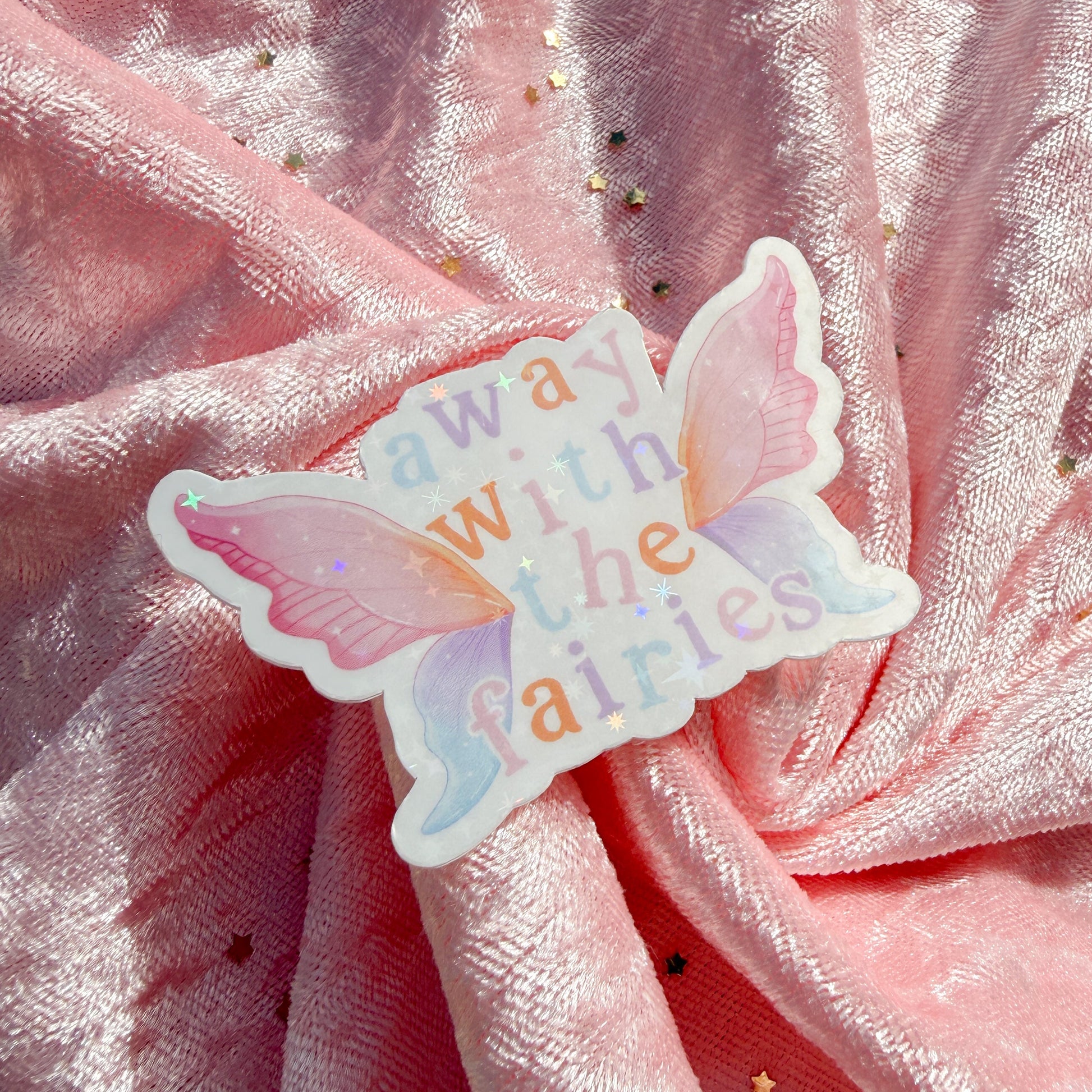 A butterfly-shaped sticker with the words 'away with the fairies' in pastel colors, placed on a draped pink glittery fabric background.