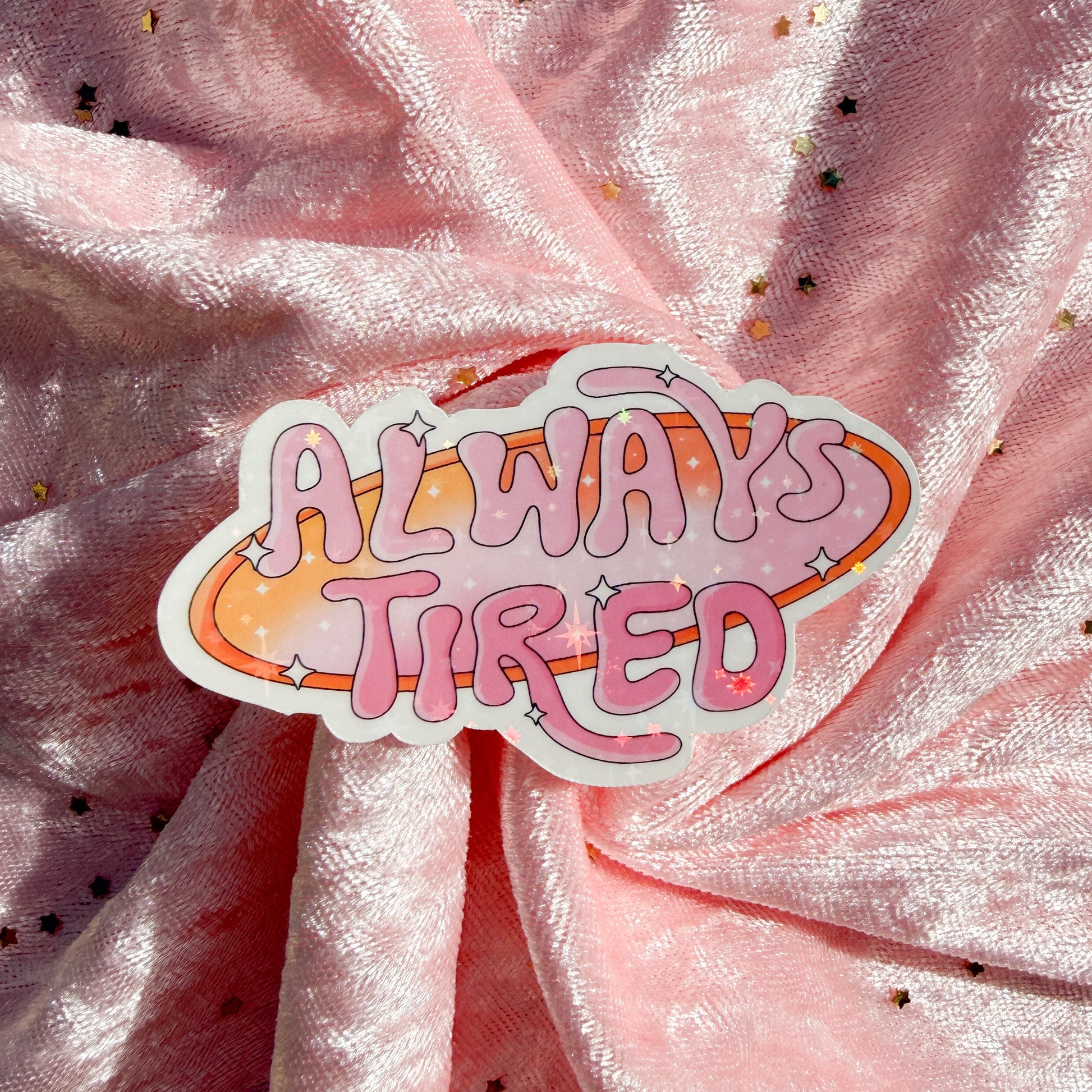 A die-cut sticker with the phrase 'ALWAYS TIRED' in pink bubble letters over a watercolor-style orange and pink space ring, placed on draped pink fabric with small gold star confetti.