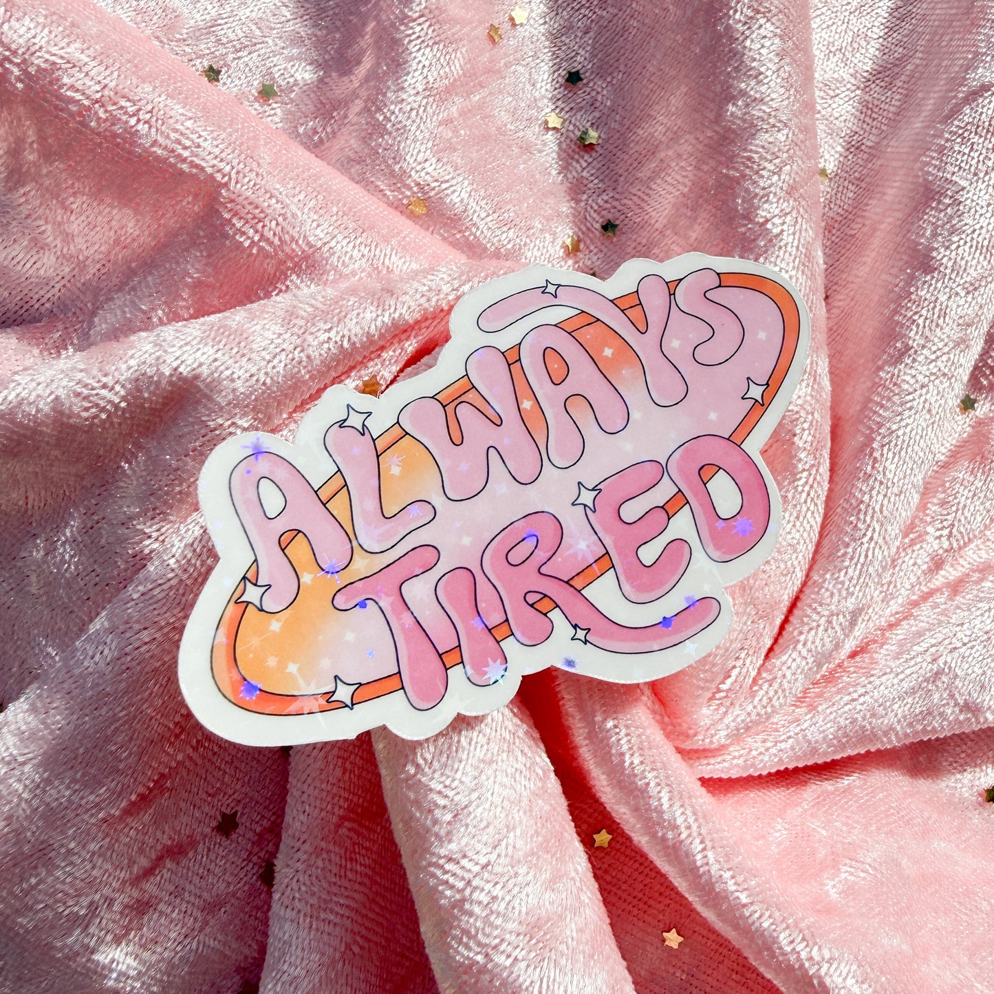 A die-cut sticker with the phrase 'ALWAYS TIRED' in pink bubble letters over an orange and pink cloud-like shape with small star accents, placed on draped pink glitter fabric.