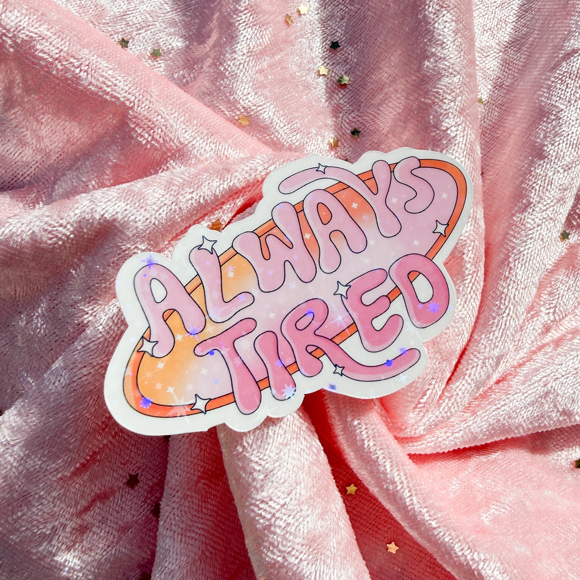 A die-cut sticker with the phrase 'ALWAYS TIRED' in pink bubble letters over an orange and pink cloud-like shape with small star accents, placed on draped pink glitter fabric.