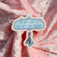A die-cut sticker featuring a light blue sign that reads 'ON MY WAY TO THE BOY AQUARIUM' with small white flowers and an illustration of ice skates hanging below, placed on pink glittery fabric.