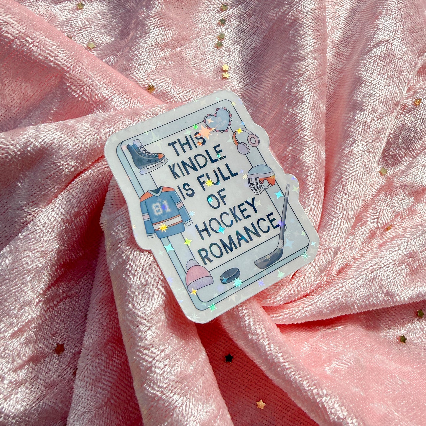 A die-cut sticker with a hockey theme and the text 'THIS KINDLE IS FULL OF HOCKEY ROMANCE' pinned onto a draped pink fabric background.