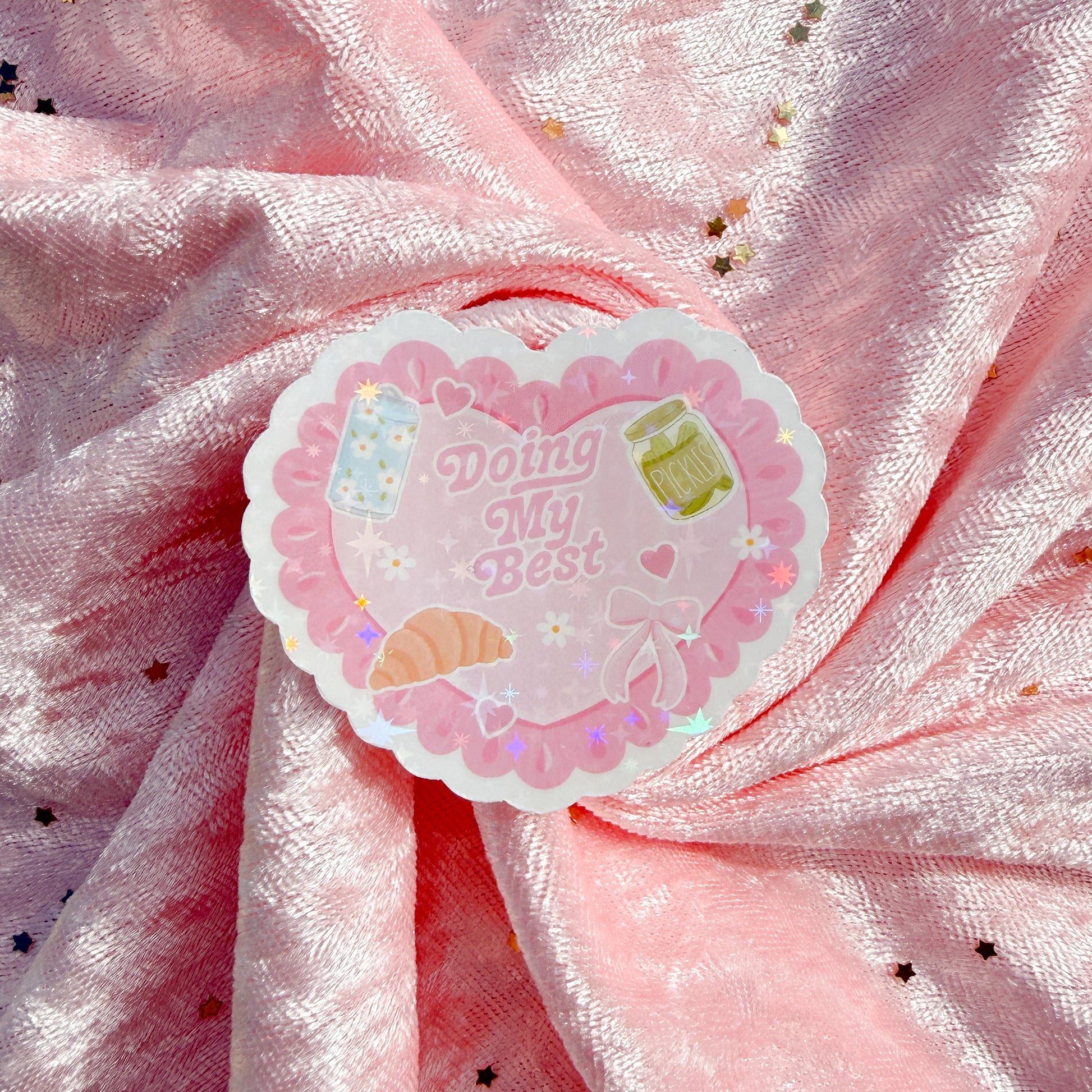 A heart-shaped sticker with a pink scalloped border and the text 'Doing My Best' in script, placed on a shiny pink fabric background with scattered gold star confetti.