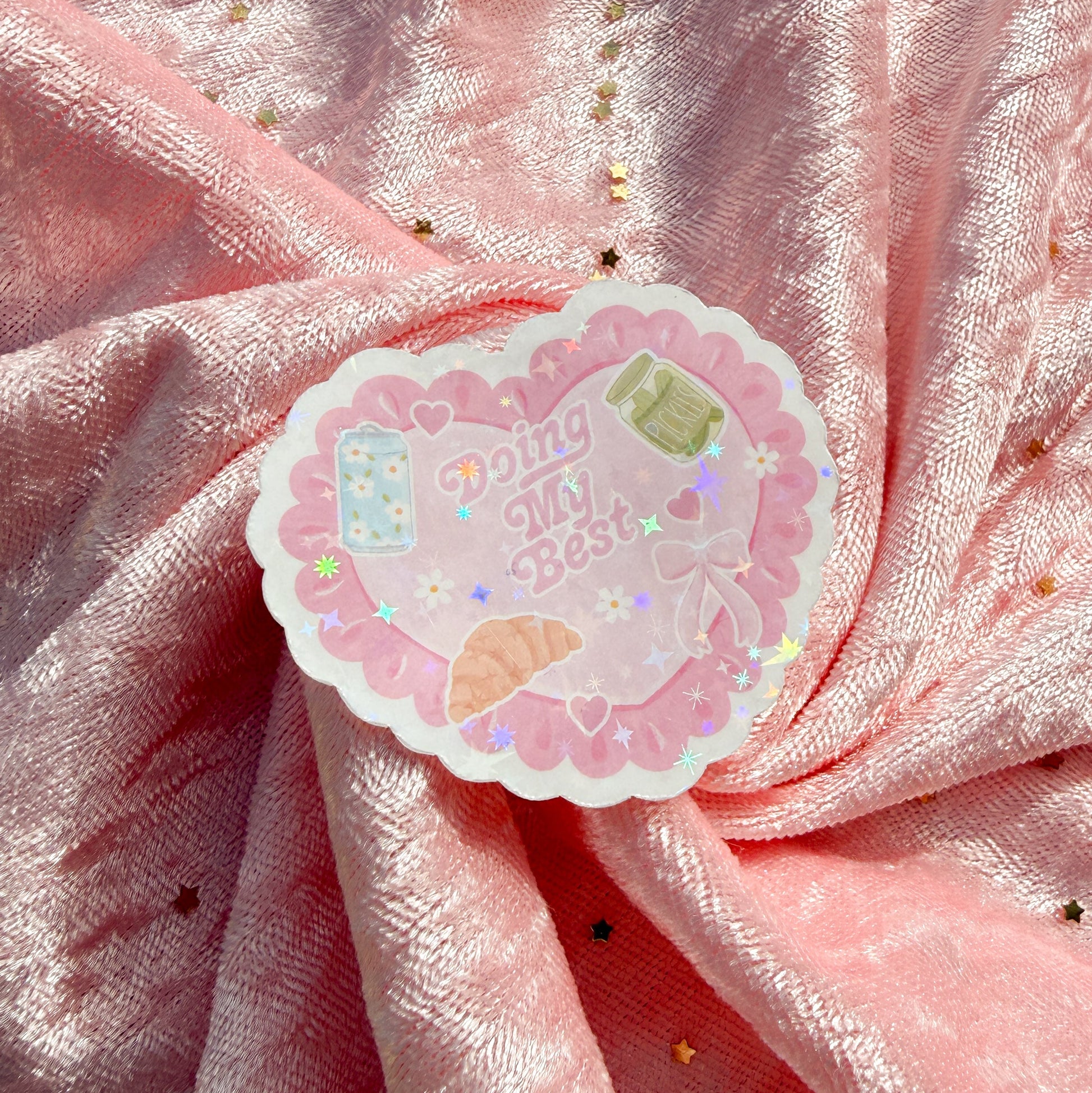 A scalloped die-cut sticker with the text 'Doing My Best' in pink lettering, surrounded by cute illustrations and small holographic star accents, placed on a draped pink glittery fabric background.