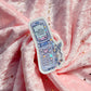 A die-cut sticker featuring an illustration of a retro flip phone with pastel blue and lavender colors, placed on a draped pink fabric background with small gold star accents.