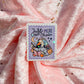 A rectangular sticker with scalloped edges featuring a cute cartoon duck reading under a crescent moon and stars, with the text 'Just MORE ONE Chapter' on a lavender background. The sticker is placed on draped pink fabric with scattered gold star confetti.