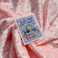A rectangular sticker with scalloped edges featuring a cute duck illustration and the text 'Just More One Chapter' lies on draped pink glitter fabric.