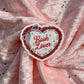 A heart-shaped sticker with the words 'Book Lover' in red script, surrounded by a red and white stitched-style border, placed on a pink glittery fabric background.