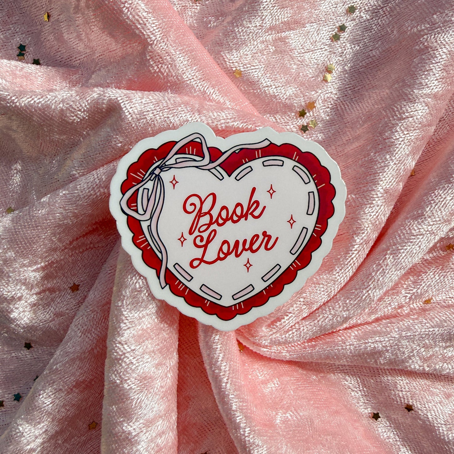 A heart-shaped sticker with the words 'Book Lover' in red script, surrounded by a red and white stitched-style border, placed on a pink glittery fabric background.