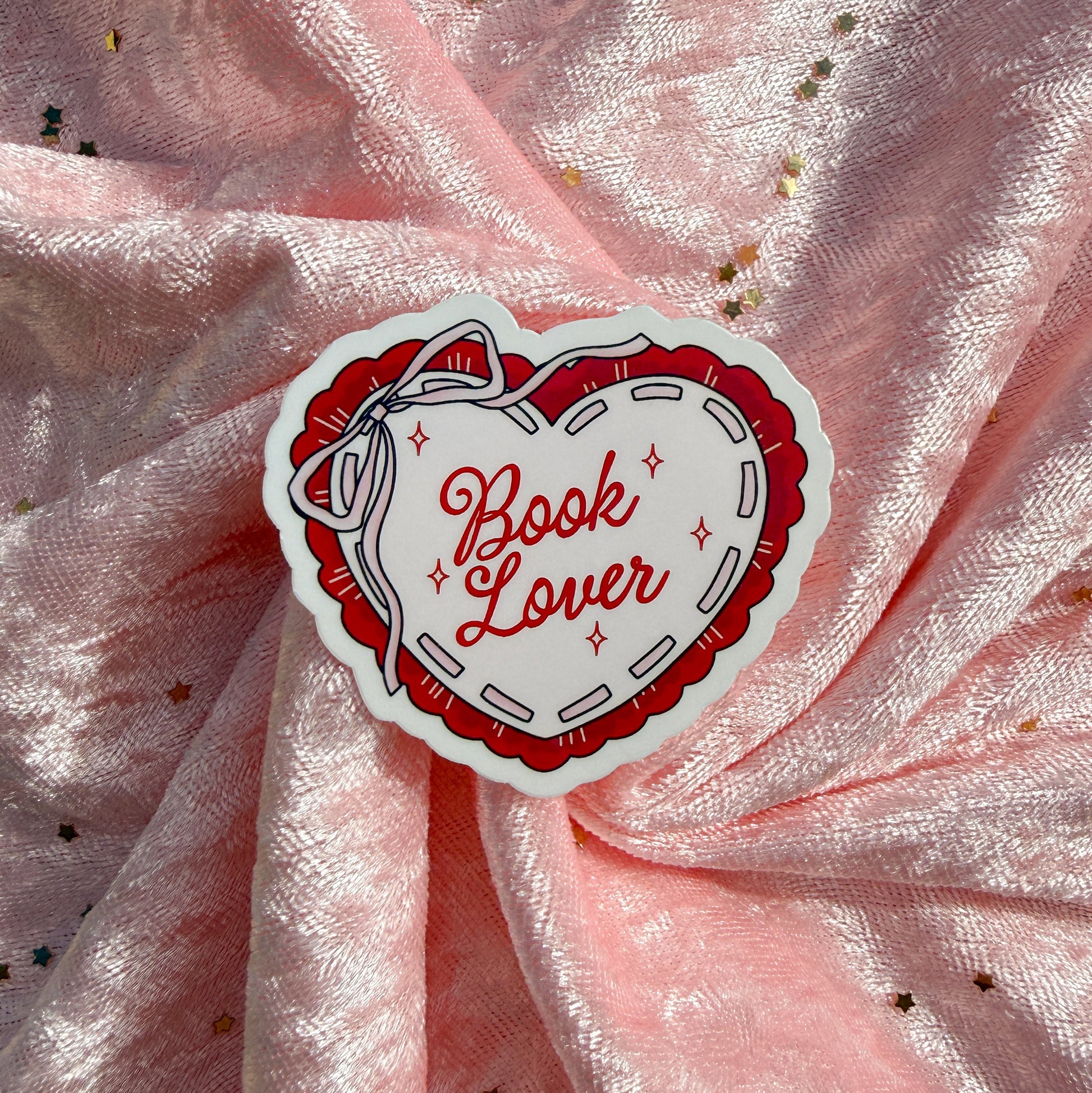 A heart-shaped sticker with the words 'Book Lover' in red script, surrounded by a red and white stitched-style border, placed on a pink glittery fabric background.