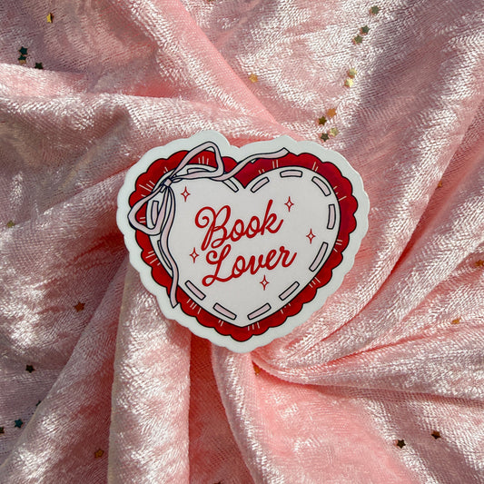 A heart-shaped sticker with the words 'Book Lover' in red script, surrounded by a red and white stitched-style border, placed on a pink glittery fabric background.