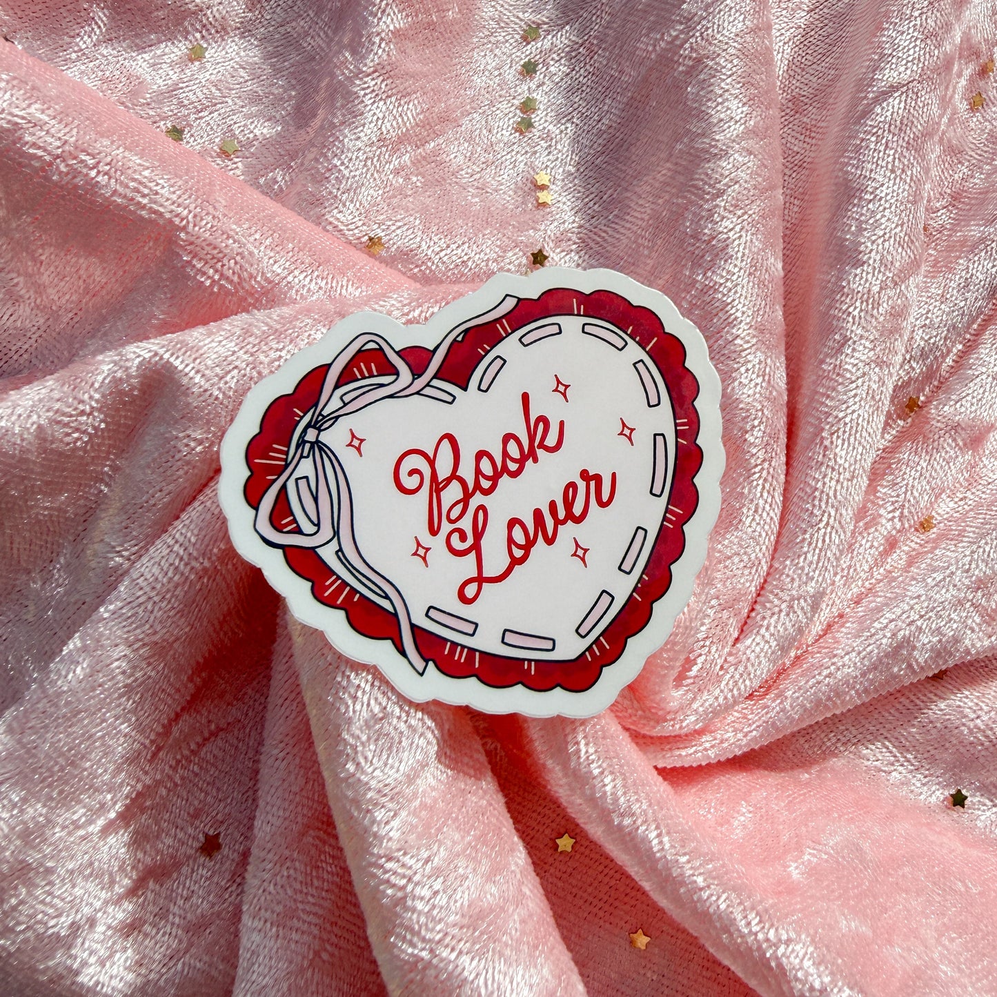 A heart-shaped sticker with the words 'Book Lover' in red script inside a red stitched border, placed on a pink satin fabric background with small gold star accents.
