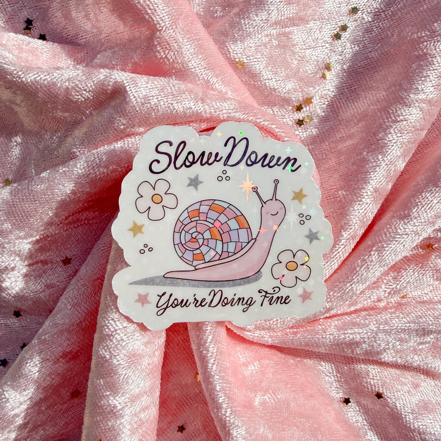 A die-cut sticker featuring a cute pink snail with a mosaic shell and the text 'Slow Down You're Doing Fine', placed on a shiny pink fabric background with gold star confetti.