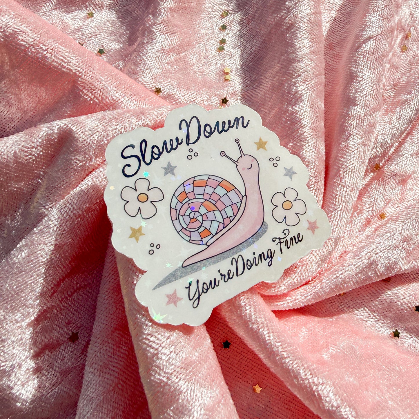 A die-cut sticker featuring a pink snail and the text 'Slow Down You're Doing Fine' on a pink glittery fabric background.