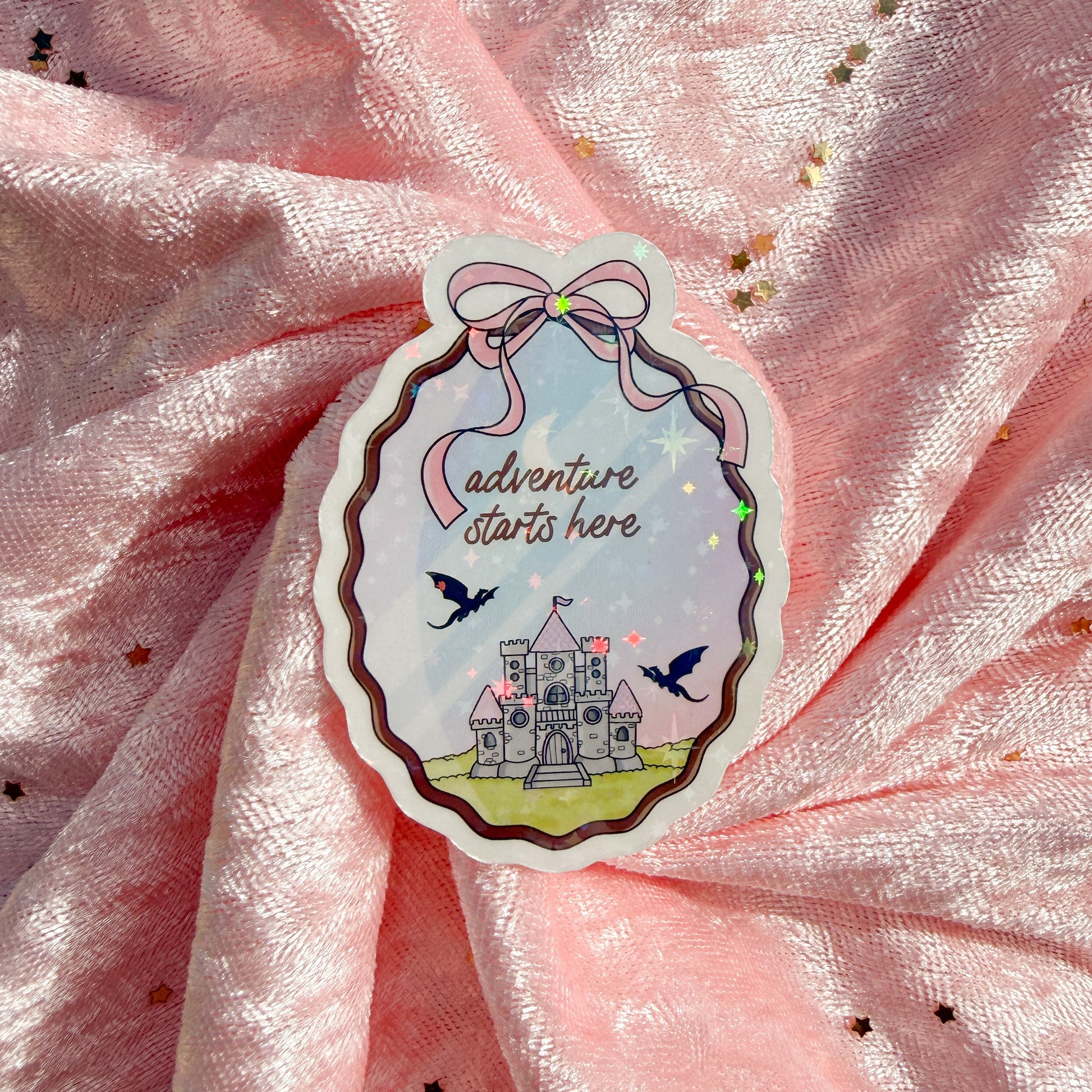 A die-cut sticker featuring a whimsical castle illustration with the text 'Adventure starts here' and small birds, placed on a pink glittery fabric background.