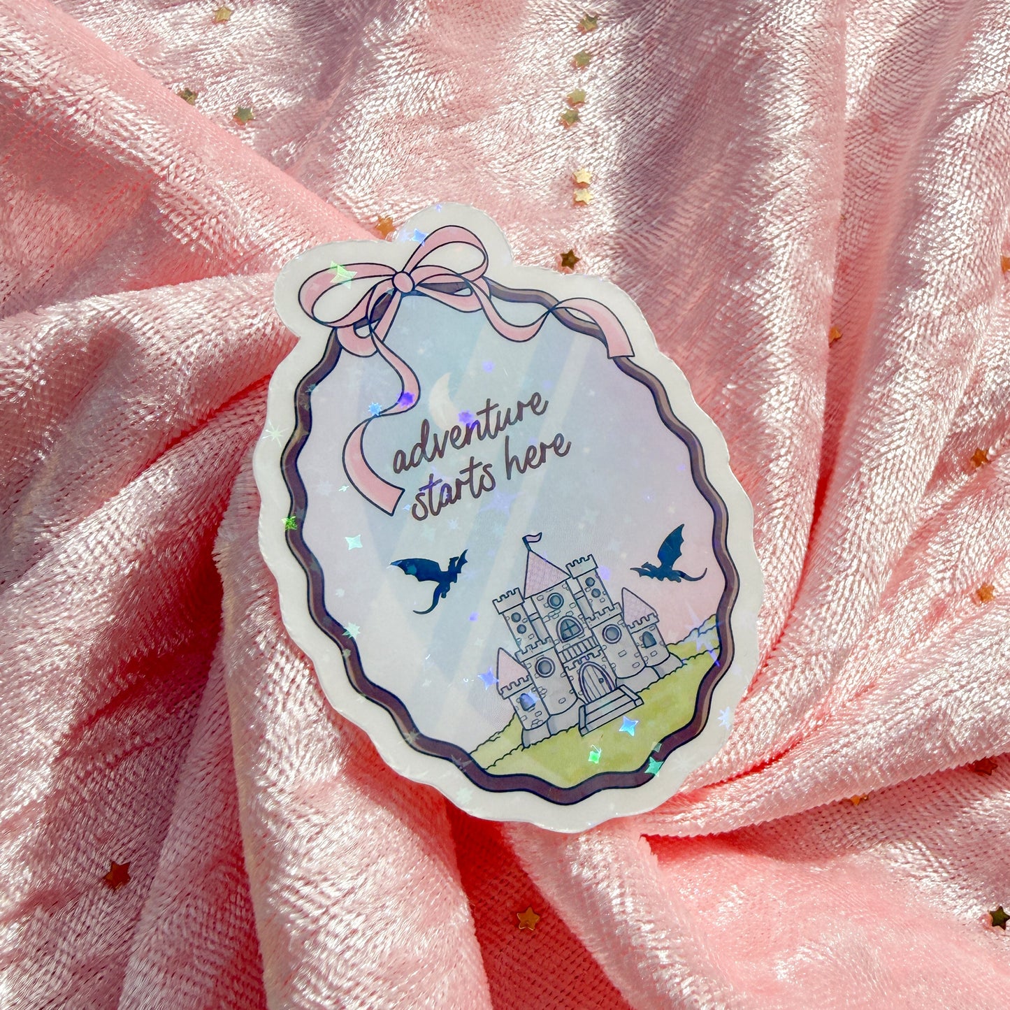A die-cut sticker with a whimsical castle illustration and the text 'Adventure starts here' attached to a pink fabric backdrop with gold sequins.