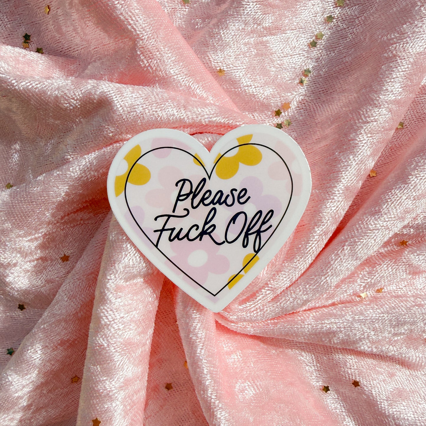 A heart-shaped sticker with the text 'Please Fuck Off' in black script, decorated with yellow dots and a black heart outline, shown on a draped light pink fabric background with small gold star details.