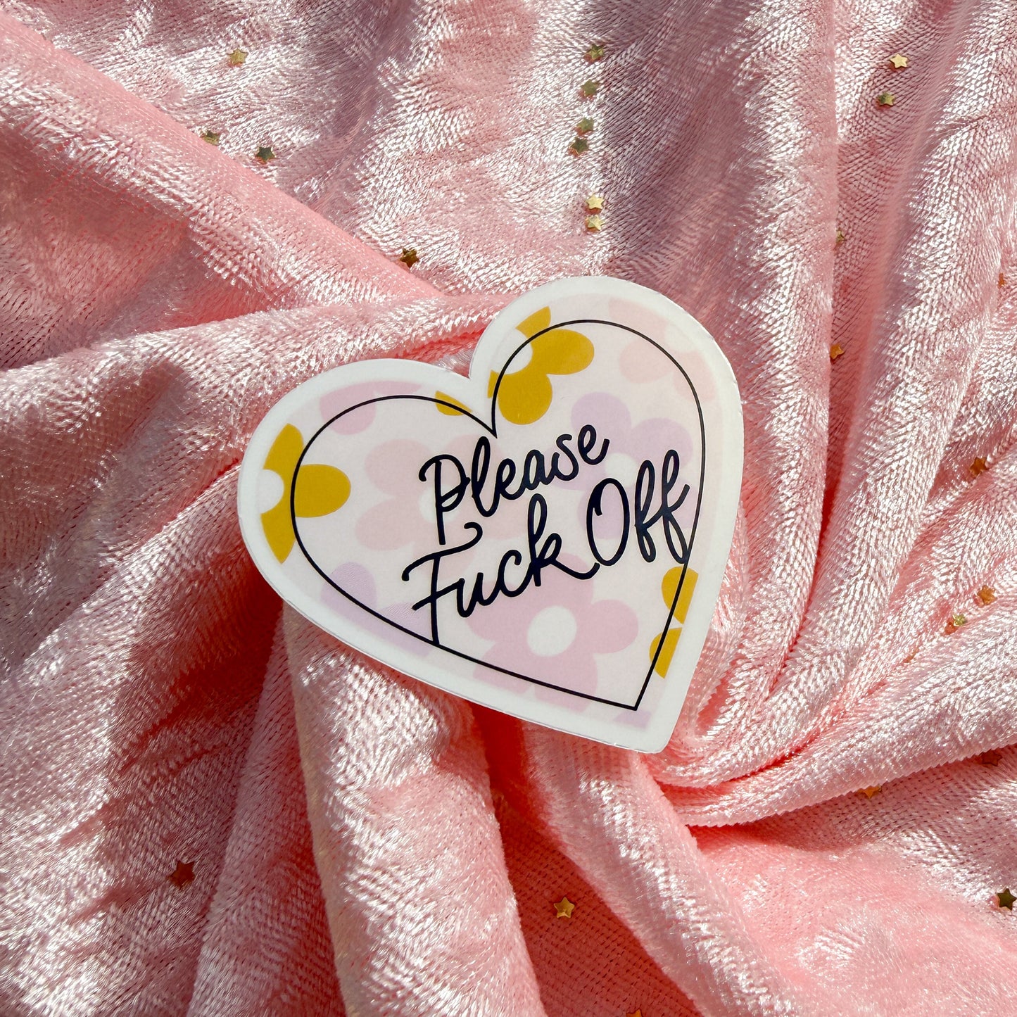 A heart-shaped sticker with the text 'Please Fuck Off' in black script, placed on a shiny pink fabric background with gold glitter.