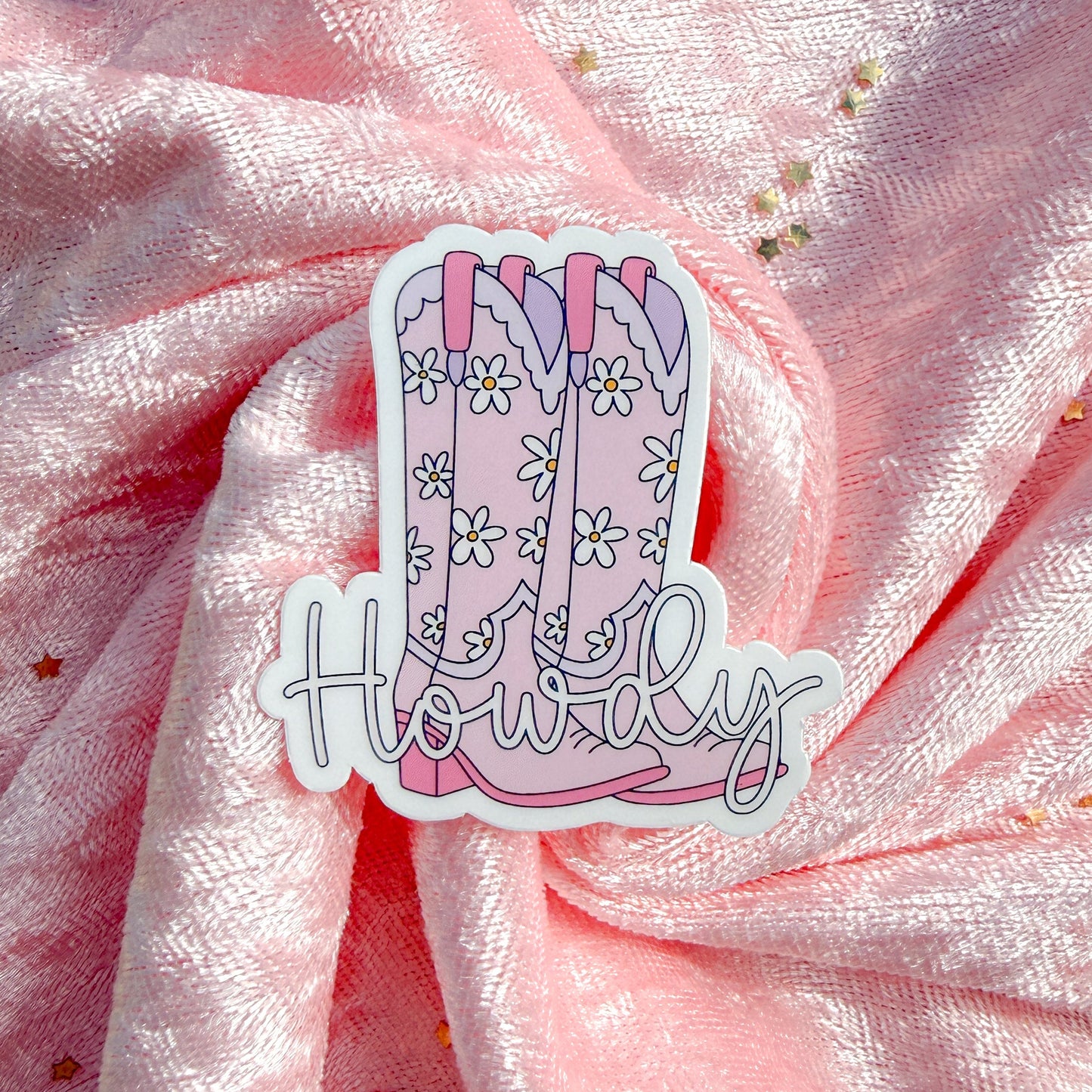 A die-cut sticker featuring a pair of pink cowboy boots with white daisy patterns and the word 'Howdy' in script, placed on a pink glittery fabric background.