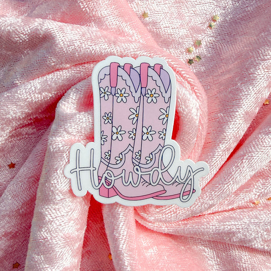 A die-cut sticker featuring a pair of pink cowboy boots with white daisy patterns and the word 'Howdy' in script, placed on a pink glittery fabric background.