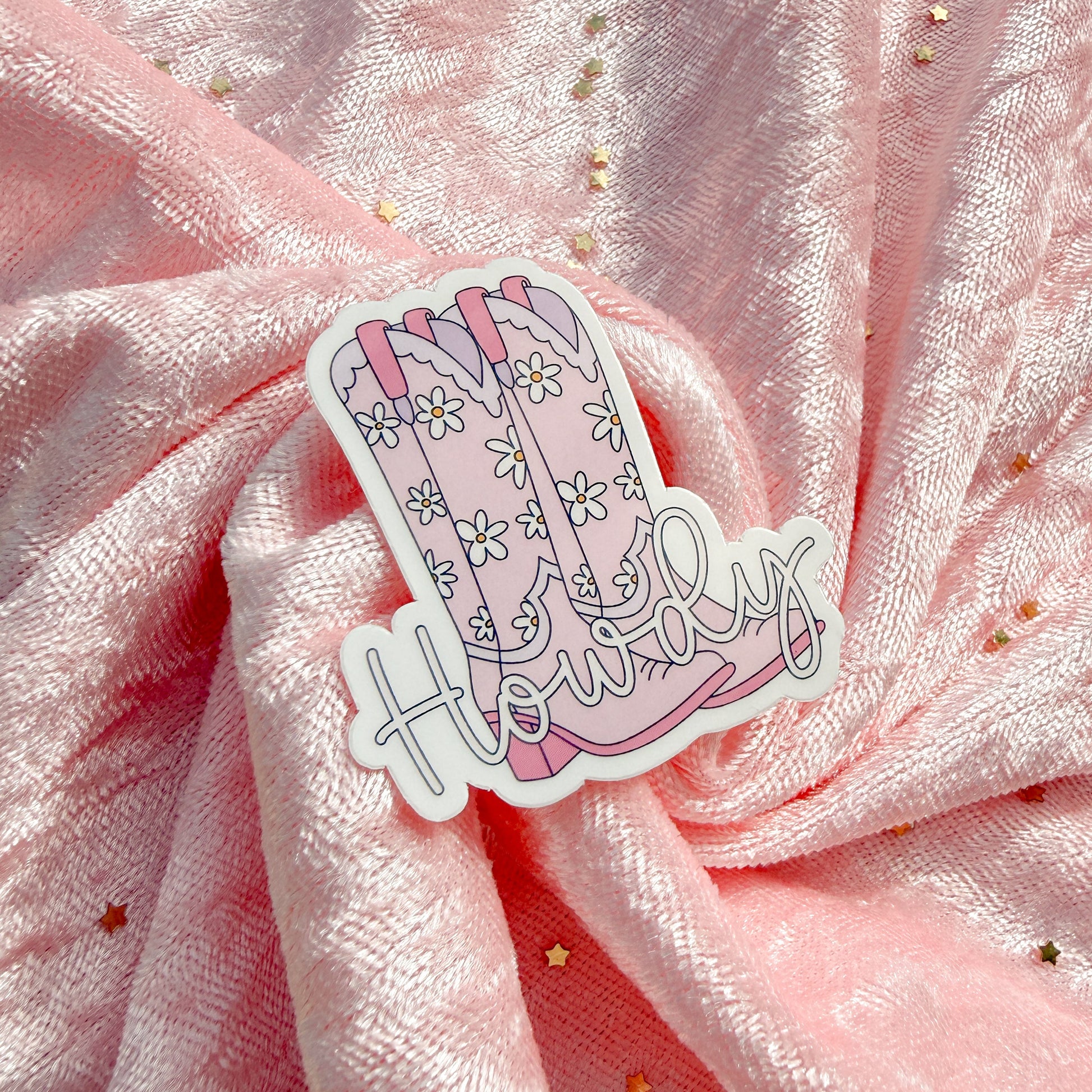 A die-cut sticker featuring a pink floral boot and the word 'Howdy' in cursive, placed on draped pink fabric with small gold star accents.