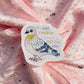 A die-cut sticker featuring an illustrated pigeon with the text 'IF I WAS A PIGEON I WOULD SHIT ON YOU' in pastel lettering, placed on a shiny pink fabric background with scattered gold star confetti.