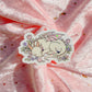 A die-cut sticker featuring a sleeping white dragon with pink wings, surrounded by small flowers and mushrooms, placed on a shiny pink fabric background with scattered gold glitter.