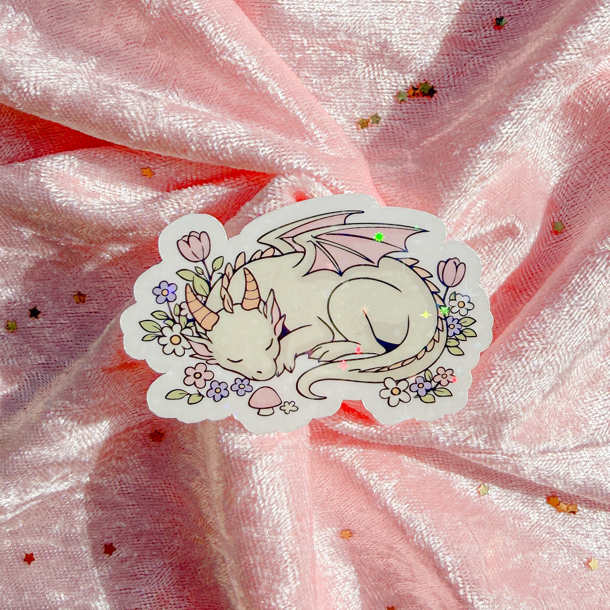 A die-cut sticker featuring a sleeping white dragon with pink wings, surrounded by small flowers and mushrooms, placed on a shiny pink fabric background with scattered gold glitter.