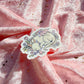 A die-cut sticker featuring a sleeping white dragon surrounded by pastel flowers, placed on a draped pink fabric background with scattered gold glitter.