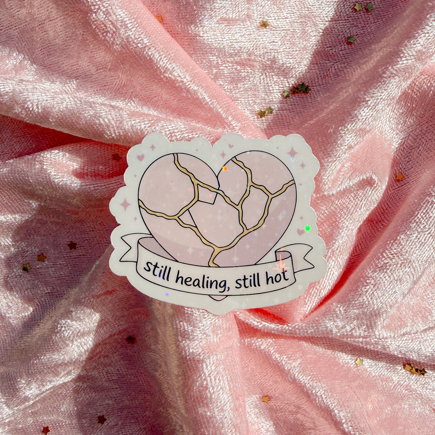 A die-cut sticker featuring a cracked pink heart with gold lines and the text 'still healing, still hot' on a ribbon banner, placed on a crinkled pink fabric background with scattered gold glitter.