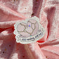 A die-cut sticker featuring a cracked pastel pink heart with the text 'still healing, still hot' on a ribbon banner, placed on draped pink fabric with scattered gold glitter.