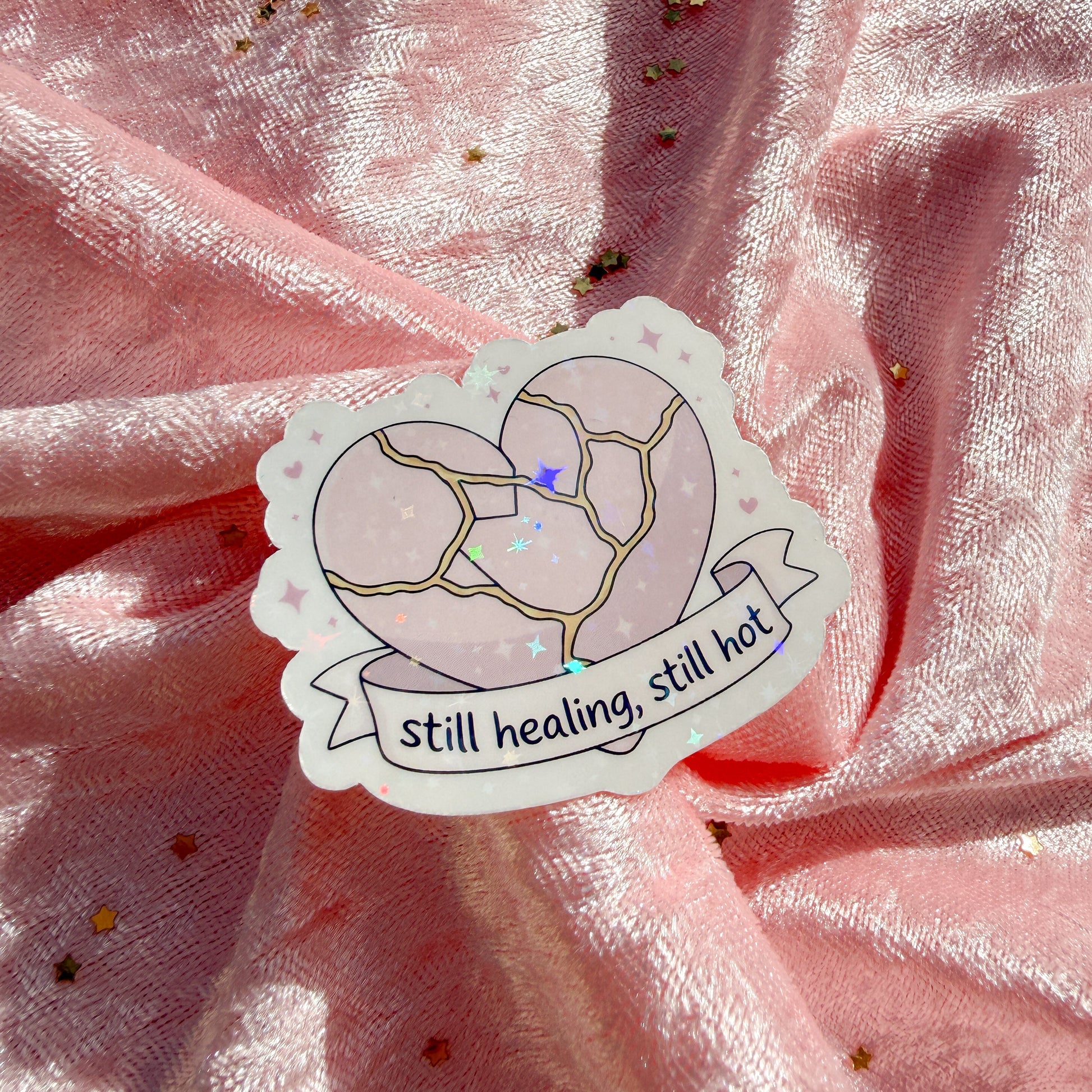 A die-cut sticker featuring a cracked pastel pink heart with the text 'still healing, still hot' on a ribbon banner, placed on draped pink fabric with scattered gold glitter.